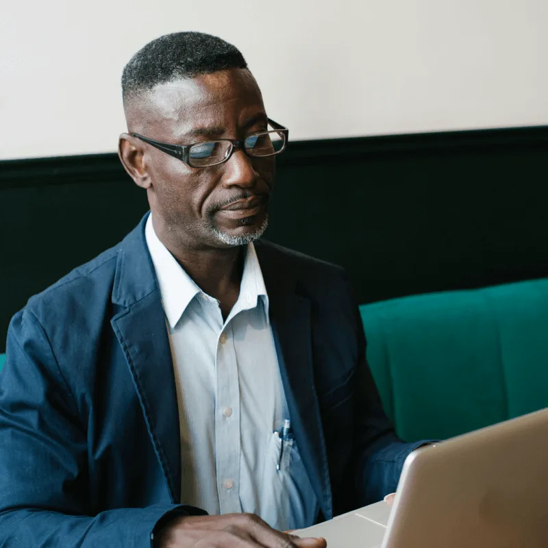 A man in casual business wear sits on a green sofa looking at his laptop